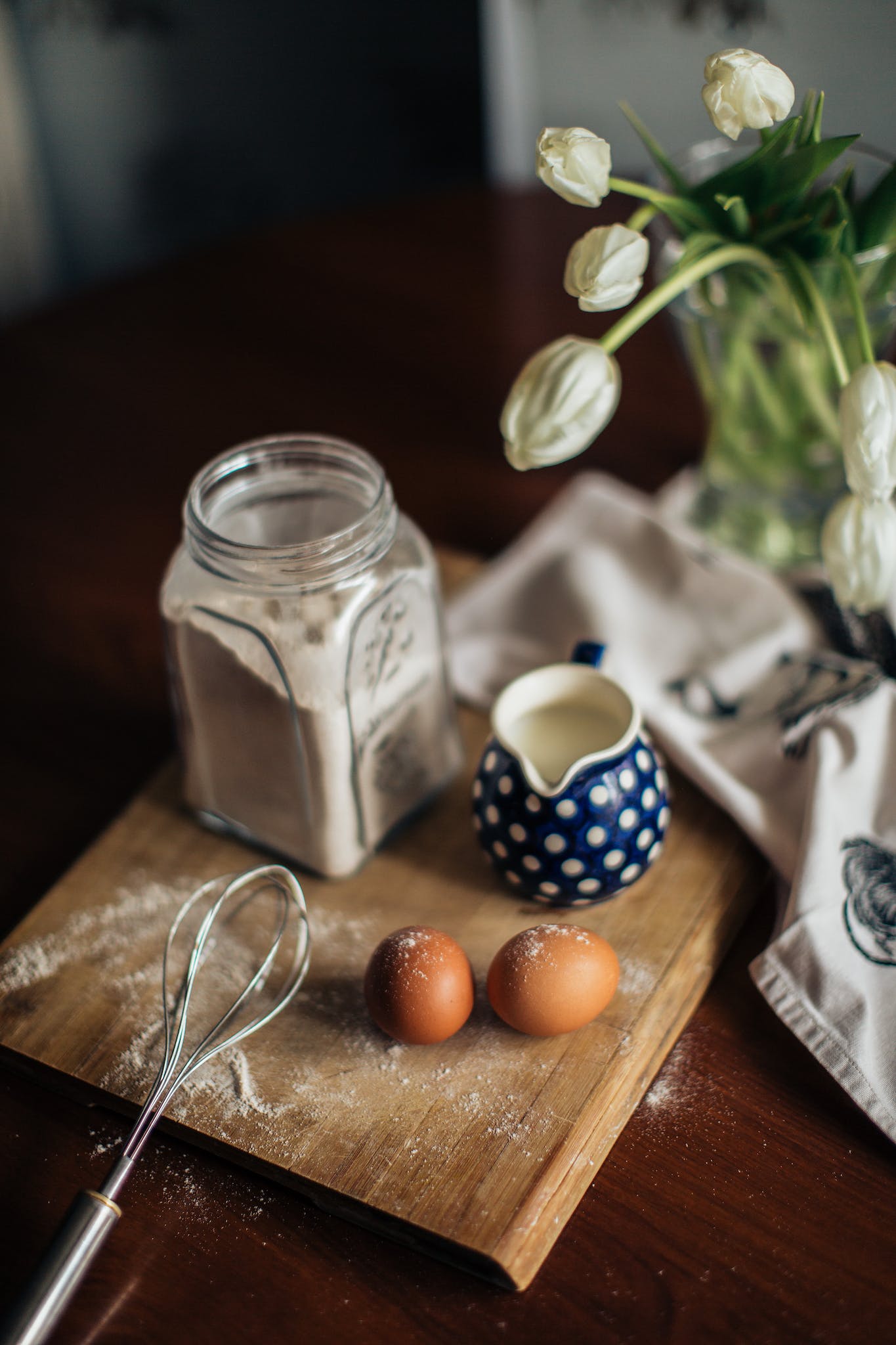 Recettes Air Fryer Du dessus du vase avec des fleurs blanches placées près des œufs de lait frais et du pot en verre avec de la farine sur une planche à découper en bois dans la cuisine